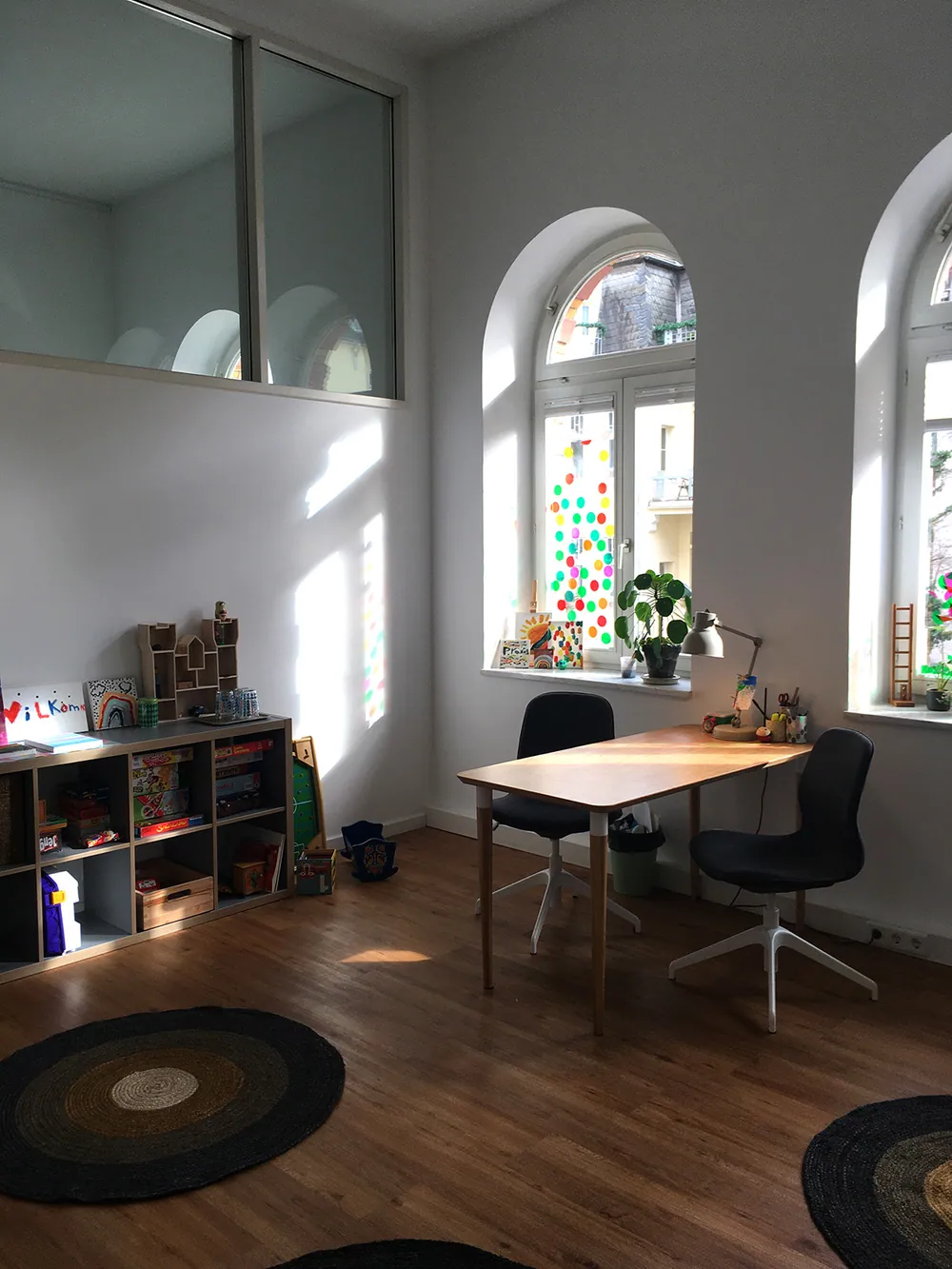 Heller Praxisraum mit hohem Altbaufenster, Spielregal und bunter Fensterdeko.